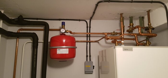 Rött expansionskärl med säkerhetsventil i källare, omgiven av kopparrör och isolerade rör, del av bergvärmeinstallation.