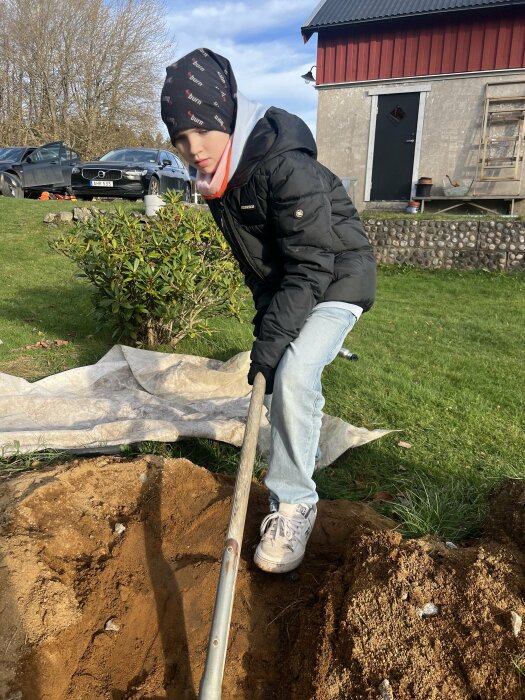 Barn i vinterkläder gräver med spade i trädgården framför ett hus. Två bilar syns i bakgrunden.