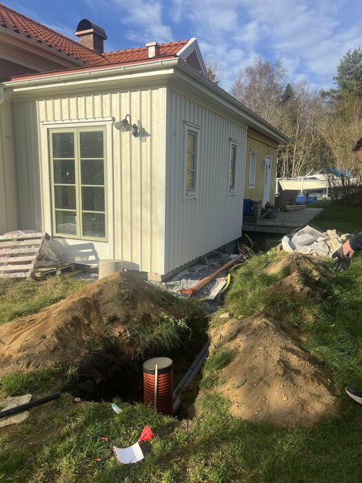 Fasadbyte och fönsterdörrsinstallation på hus; utgrävning för rörarbete i förgrunden, vårdag.