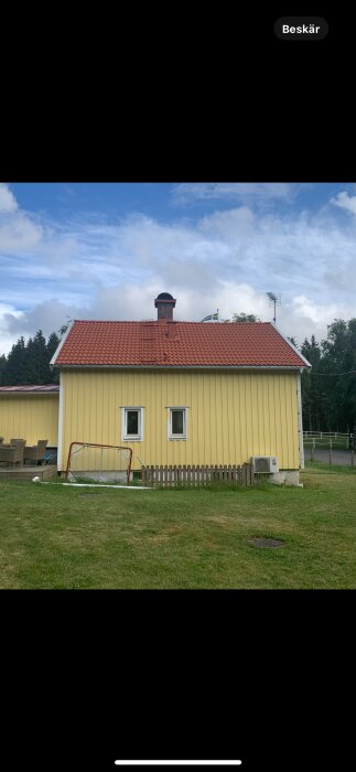 Gult hus med röd tegeltak, två fönster, närliggande staket och målnät på gräsmatta, omgiven av skog.