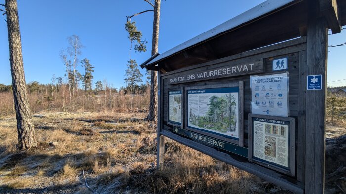 Informationsskylt vid Svartdalens naturreservat med text och bilder om skogen. Bakom syns ett skogsområde under en klarblå himmel.