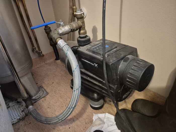 Svart Grundfos vattenpump med slangar och ventiler installerad i ett hus, inklämd mellan andra VVS-komponenter på ett golv.