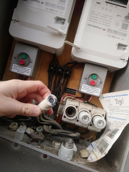 En trasig säkring hålls av en hand framför ett elskåp med två elmätare märkta Vattenfall och flera kablar och anslutningar synliga.