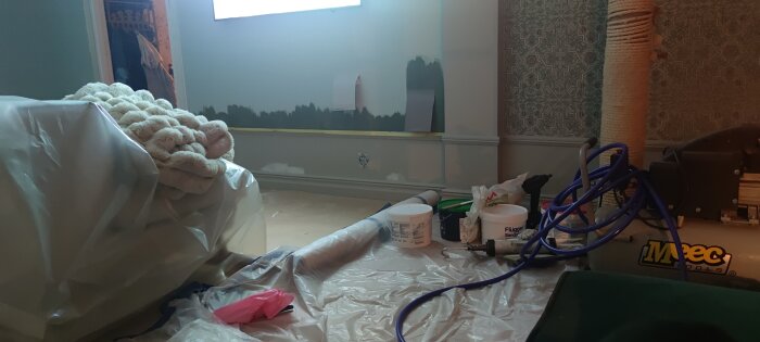 Renovering av vardagsrum med plast över möbler, målarfärg på väggen och målarverktyg på golvet. Provmålning av tv-vägg.