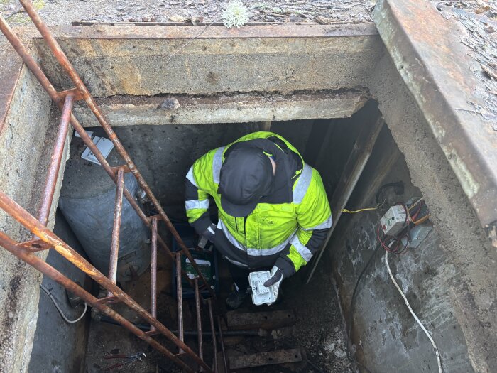 Person i skyddskläder undersöker betongrum under mark, rostig stege leder ner, väggar visar avflagnad puts och exponerade kablar.