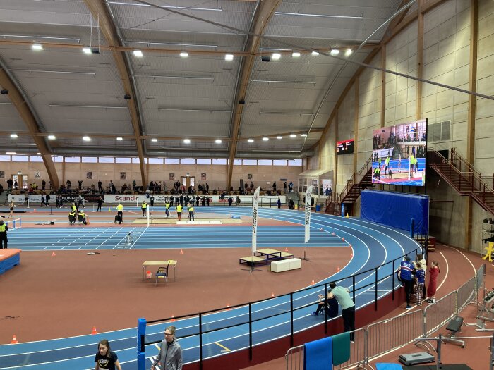 Inomhus friidrottsarena med blå löparbanor, människor i gula västar, ett höjdhoppområde och en stor skärm som visar en sporthändelse.