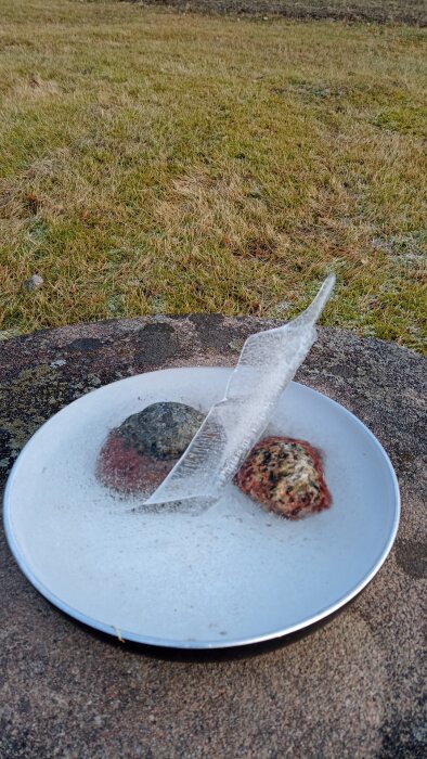 Naturligt bildad iskonst i skål, med en cirka 15 cm hög, lutande ispelare på fryst vatten. Skålen står på en sten.