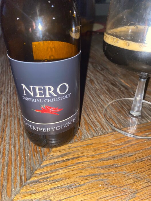 Flaska med etikett "Nero Imperial Chili Stout" från Imperiebryggeriet, stående på ett träbord bredvid ett halvtomt glas stout.