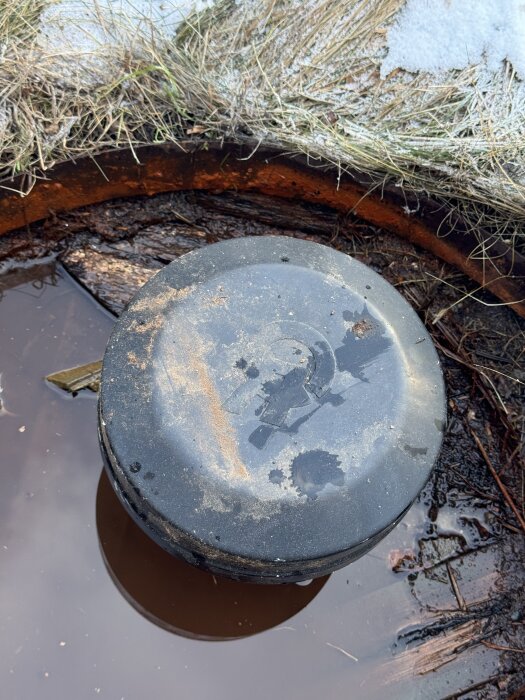 Bilden visar en svart borrtopp med rostfläckar och vattenpöl runt omkring, omgiven av växtlighet och lite snö.