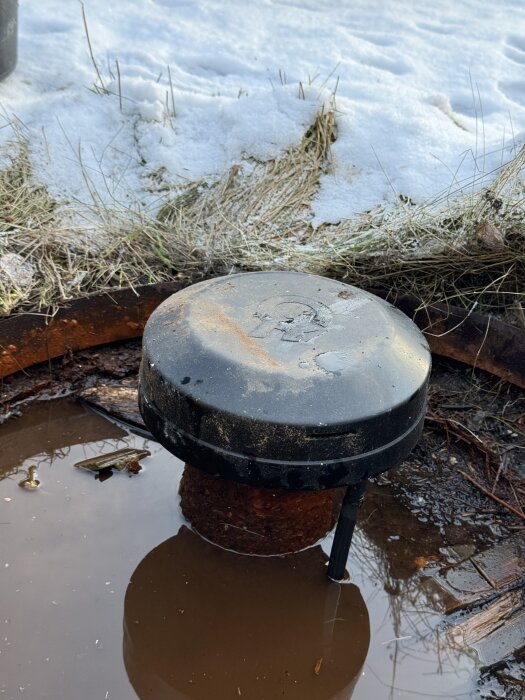 Borrtopp på djupborrad brunn med vattenpöl runt på frusen mark, eventuellt problem med läckage.