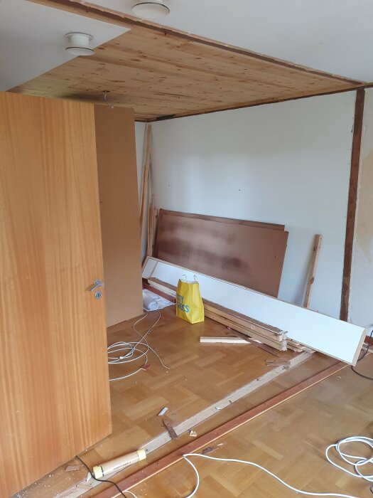 Renoveringsprojekt i sovrum med trägolv, verktyg, träpaneler, kablar och en öppen dörr.