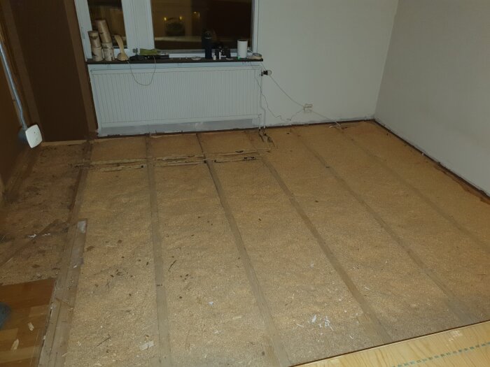 Ett sovrum under renovering, golvet är avlägsnat för att visa isoleringen och reglarna. Ett fönster med några föremål på fönsterbrädan syns.