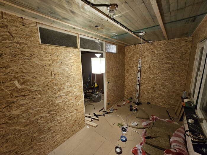 Renovering av sovrum med träpanelade väggar och oinreddt tak; verktyg och material ligger spridda på golvet.
