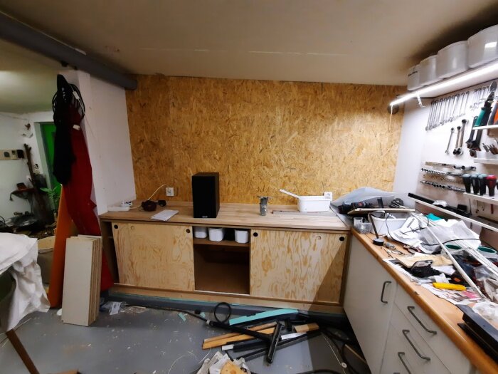 Verkstad med plywoodväggar, arbetsbänk och verktyg, inklusive skruvmejslar och dosor, med plywoodmaterial på golvet.
