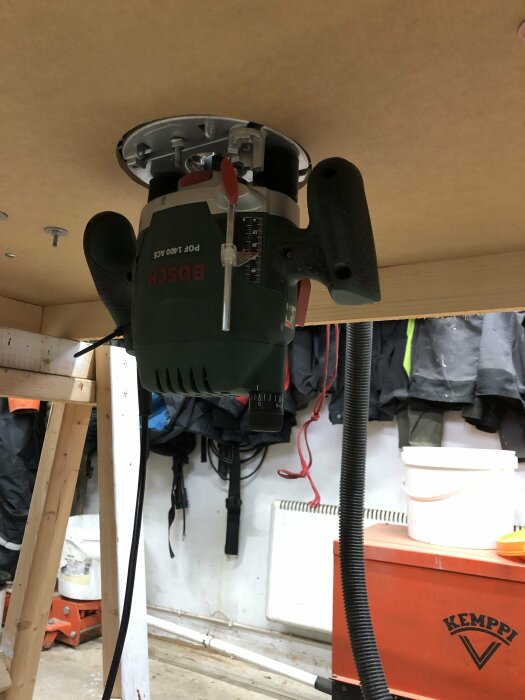 Bilden visar en Bosch POF 1400 ACE router monterad upp och ner på ett arbetsbord i ett garage, omgiven av verktyg och utrustning.