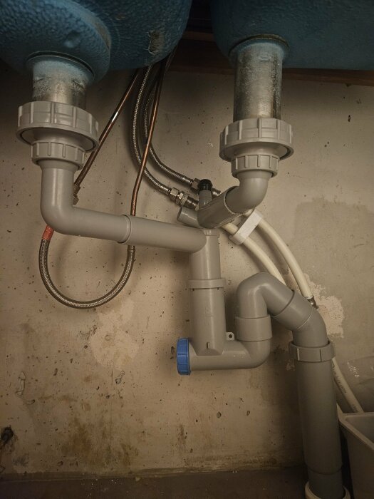 Vattenrör och avlopp under diskho i 50-talslägenhet, redo för eventuell diskmaskinsinstallation. Gråa och metalliska rör mot en betongvägg.