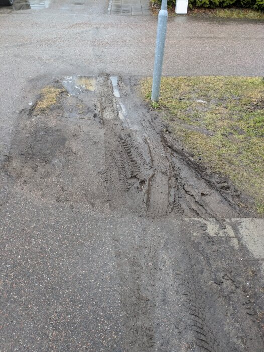 Lerig uppfart med djupa spår och pölar vatten, bredvid en gräsplätt och stålstolpe. Behöver en tillfällig beläggningslösning.