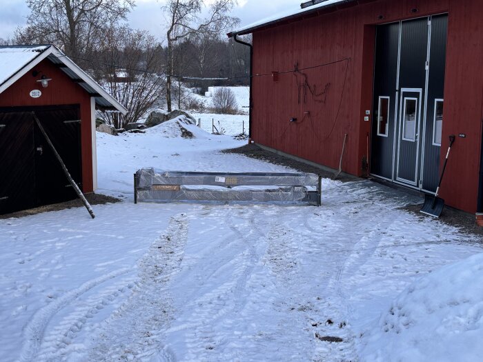 En billyft står inpackad med metallställningsben framför en röd byggnad i snöig omgivning, med en snöskyffel lutad mot väggen.