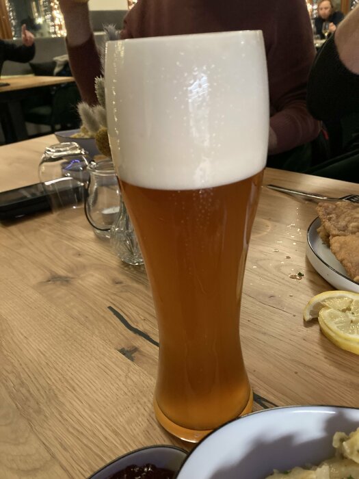 En hög glas öl med tjockt skum står på ett träbord i en restaurangmiljö. I bakgrunden finns tallrikar och en skiva citron.