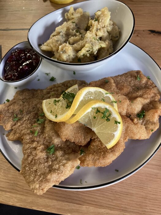 Panerad schnitzel med citronskivor, potatissallad i skål och rårörda lingon på tallrik.