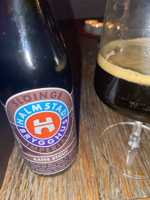 Flaska och glas med kaffe stout från Halmstads brygghus på ett bord, bakgrund med tända ljus.