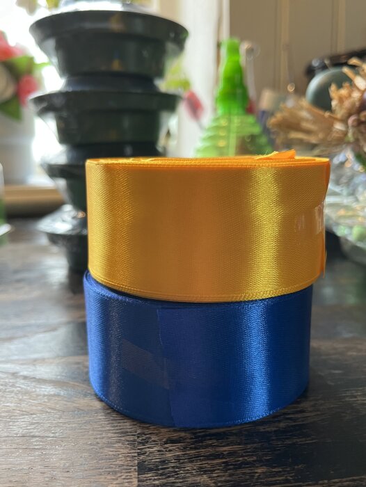 Två rullar satinband, en gul och en blå, står staplade på ett bord med suddig bakgrund av krukor och växter.