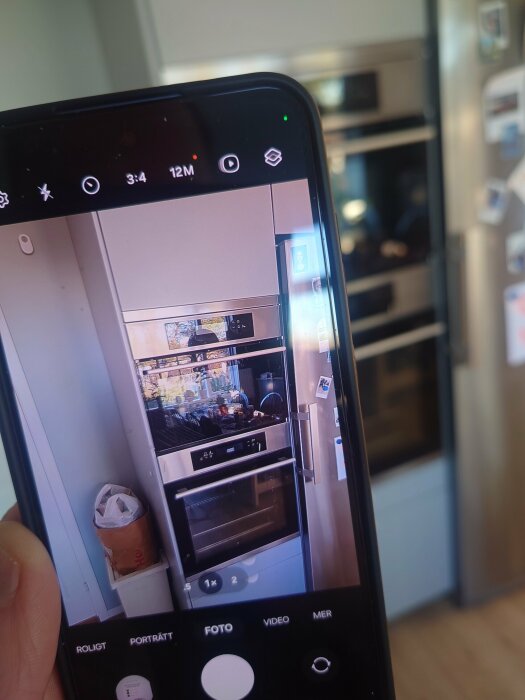 Smartphone-skärm visar foto av Ikea-kök med inbyggd ugn och mikrovågsugn och vitvaror tydliga i spegelbilden på kameran.