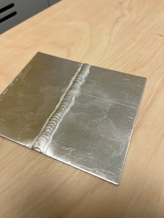 TIG-svetsad provbit i tunn aluminiumplåt på arbetsbänk, med synlig svetsfog i mitten.