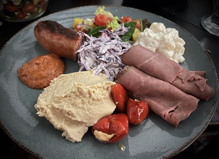 Tallrik med bratwurst, rostbiff, potatissallad, coleslaw, hummus, sallad, chili-tomatröra och ostfyllda chilis.