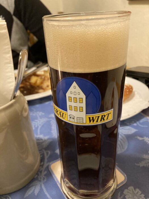 Mörkt ölglas med skummande yta, logotyp "BRÄU WIRT" på glaset, placerat på en dukad restaurangbord med servetter och tallrik i bakgrunden.