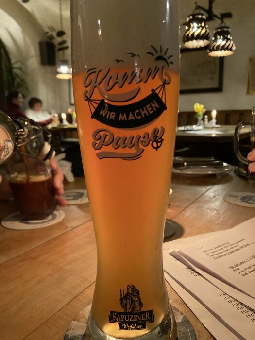 Ett högt glas med öl märkt "Kapuziner Weißbier" på ett träbord i en restaurangmiljö.