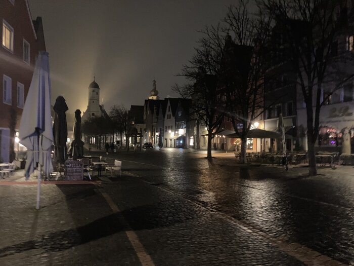 Nattvy av ett tomt torg med stenläggning, omgivande byggnader, träd med bar kvist och kyrktorn belyst i bakgrunden.