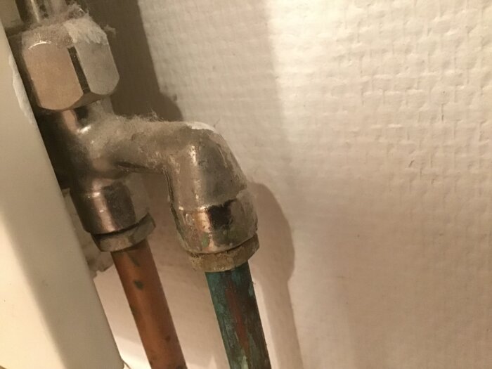 Radiatorventil med damm, ansluten till koppar- och grönt rör, installerad mot en vit vägg. Ventil från 1988 med möjligt justeringsbehov.