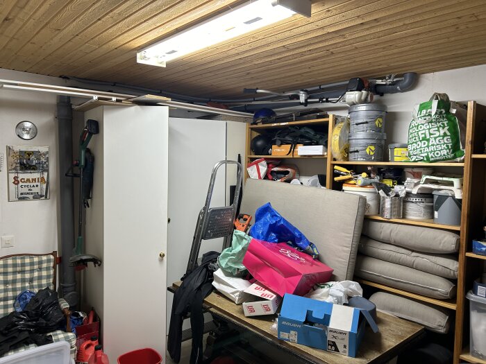 Stökigt förråd med kartonger, verktyg och diverse föremål staplade på hyllor och golv, en bänk med kuddar och påsar. Trätak med lysrör.