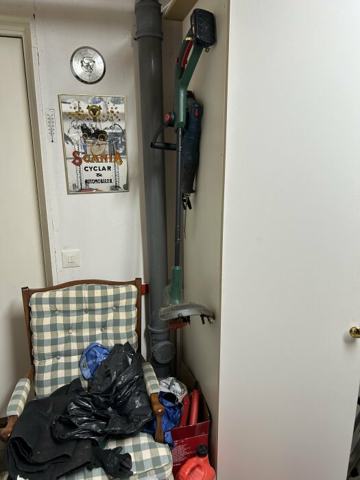 En rutig stol fylld med svarta sopsäckar bredvid en grå ventilationsrör, verktyg, en retroaffisch och en klocka på väggen.