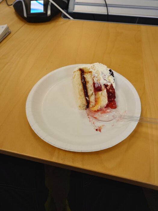 En bit tårta med grädde och sylt på en vit pappersassiett, placerad på ett träbord.