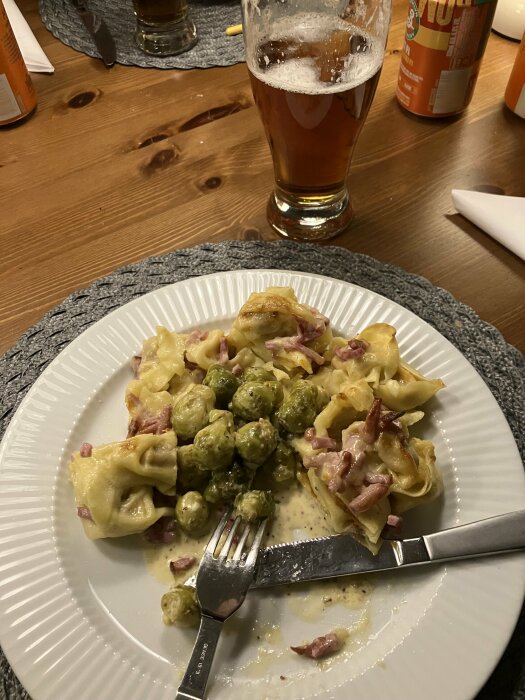 Tallrik med senapsstuvad brysselkål och pastagratäng med skinka, framför ett glas öl på ett träbord.