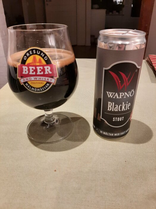 Ett glas och en burk med WAPNÖ Blackie stout, en mörk öl, på ett bord. Glaset har ett Öresund Beer and Whisky Festival-märke.