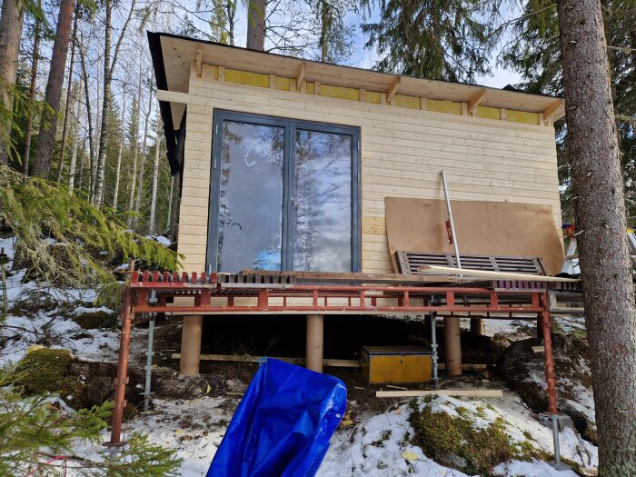 Byggnad under uppförande med nyligen installerade fönster och panel, omgiven av snötäckta träd, sedd från altandörröppningen.