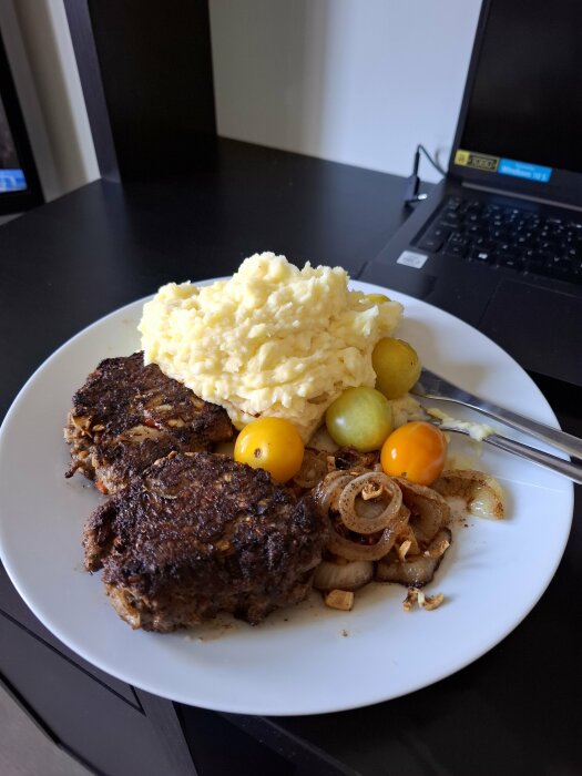 Tallrik med potatismos, pannbiffar, stekt lök och körsbärstomater på ett bord framför en laptop.