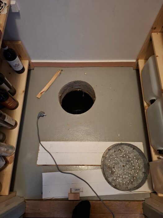 Hål i golvet i ett skafferi med vatten i botten, omgiven av trähyllor och diverse föremål, inklusive en kabel och ett perforerat metalllock.