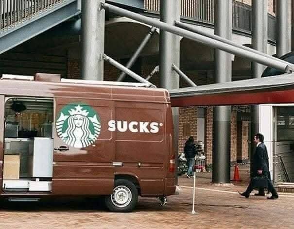 Brun bil med delvis täckt Starbucks-logga som bildar ordet "SUCKS", parkerad vid byggnad med gångbroar.