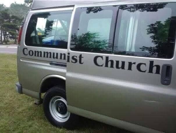 En beige minibuss med texten "Communist Church" på sidan, parkeras på gräs med träd i bakgrunden.