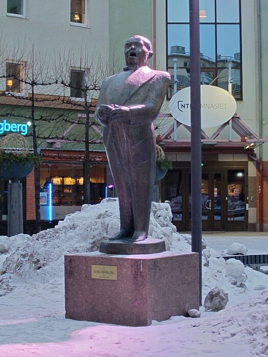Staty av en man, klädd i kavaj, stående på ett snöigt torg framför en byggnad med glasfasad och skyltar. Plaque på basen.