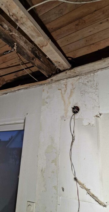 Vattenfläckar och skador på ett trätak ovanför en vägg med exponerade elkablar och fuktfläckar under en renovering.