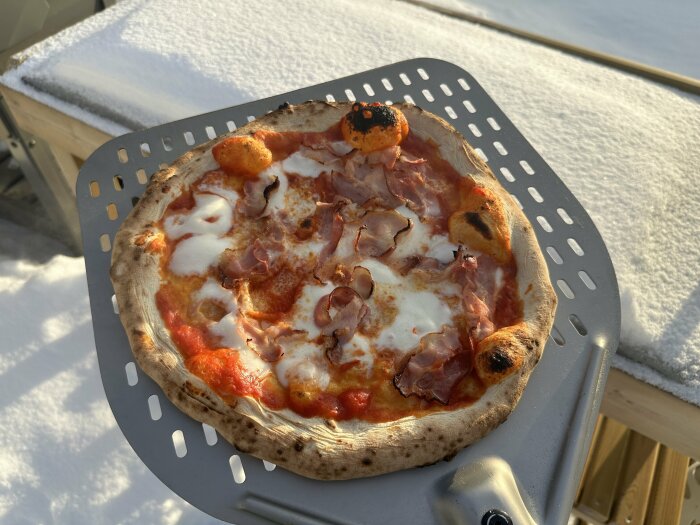 Pizza med tomatsÄs, mozzarella och skinka pÄ en pizzaspade utomhus, med snö i bakgrunden.
