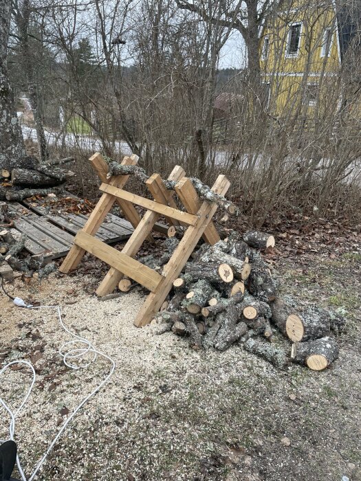 Kapat vedtravar och sågspån vid en träbock, omgiven av träd och ett gult hus i bakgrunden.