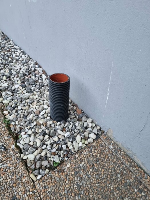 Vertikalt svart plaströr, eventuellt ett spolrör, vid grund av ett hus omgiven av småsten och grus.