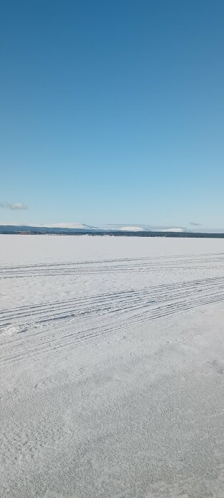 Snötäckt landskap med spår över isigt underlag under en klarblå himmel, berg synliga i fjärran.