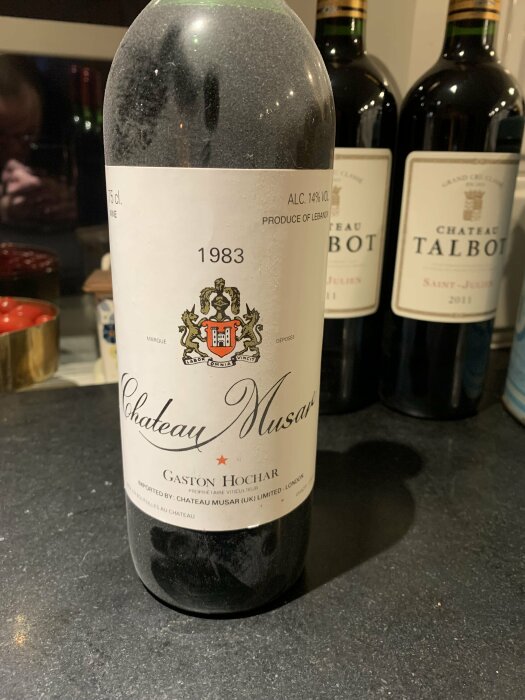 Flaska med Chateau Musar rödvin från 1983 på en bänkskiva, omgiven av andra vinflaskor i bakgrunden.